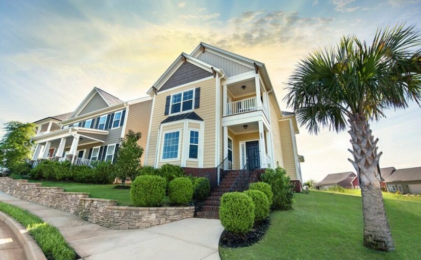 Two-story Charleston-style home with cream siding and dark roof in Greenville, South Carolina, featuring stone foundation, manicured landscaping, and palm tree in front yard