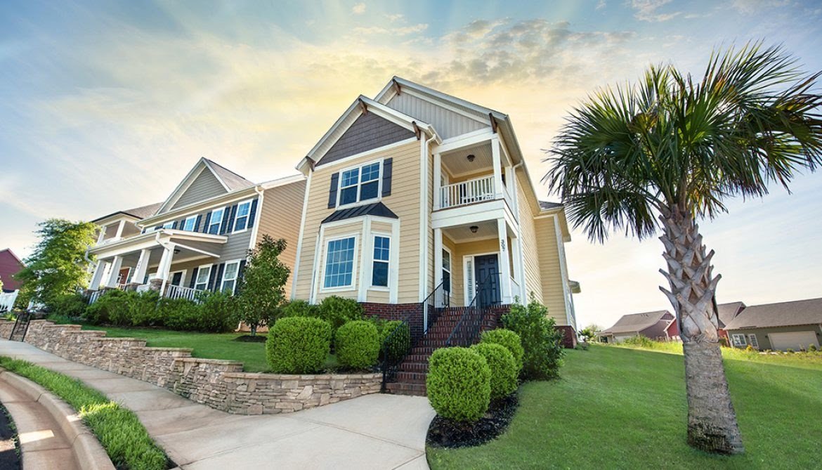 Two-story Charleston-style home with cream siding and dark roof in Greenville, South Carolina, featuring stone foundation, manicured landscaping, and palm tree in front yard