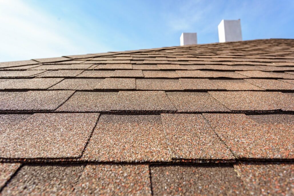 Close-up of asphalt roof shingles showing rows of brown granular shingles with chimneys visible at the peak against a blue sky
