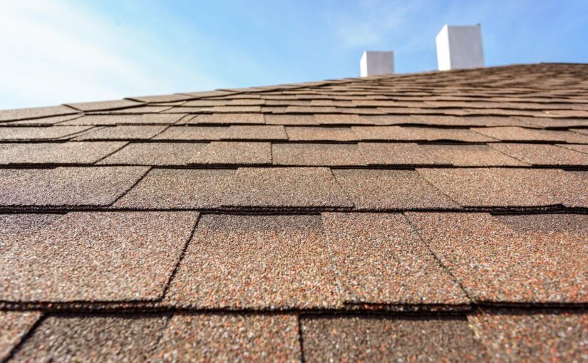 Close-up of asphalt roof shingles showing rows of brown granular shingles with chimneys visible at the peak against a blue sky