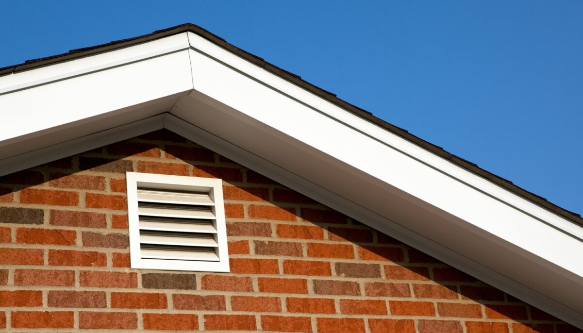 Close-up of brick gable with white soffit, fascia, and attic ventilation vent under clear blue sky