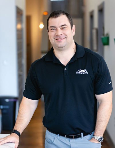 Man in black polo shirt with roofing company logo smiling in an indoor hallway setting