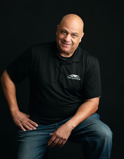 Smiling man in black polo shirt with roofing company logo, seated against dark background in professional headshot