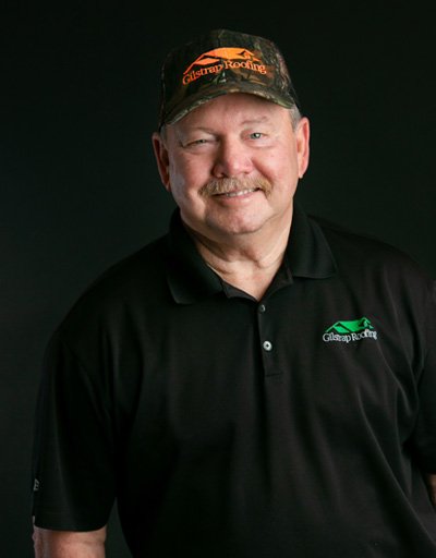 Man wearing black polo shirt with roofing company logo and camouflage cap, smiling at camera against dark background