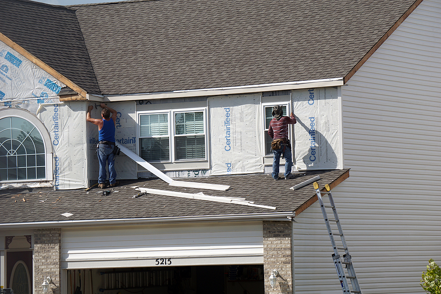 Two roofers installing new siding on a residential home during replacement project, with protective sheeting and ladder visible