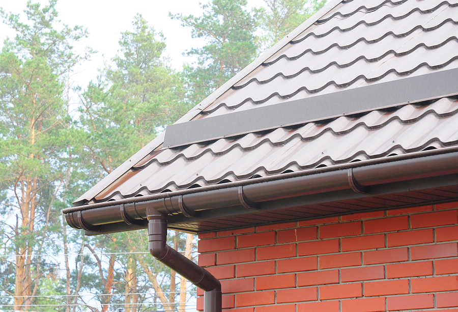 Dark metal gutter and downspout system installed on brick house with gray tile roof and pine trees in background