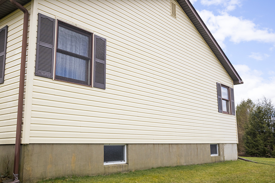 Pale yellow vinyl siding residential house with concrete foundation and dark shuttered windows in suburban setting