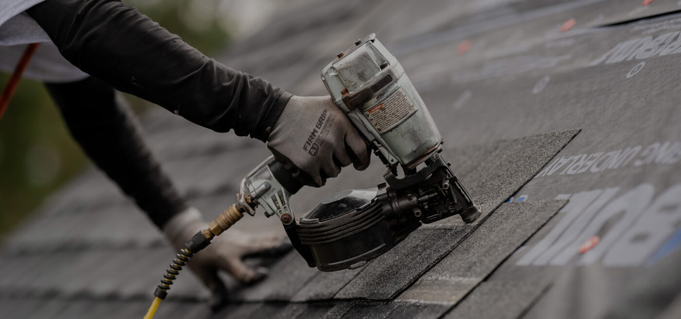 Worker in work gloves holding pneumatic nail gun against asphalt shingle roof during residential roofing installation