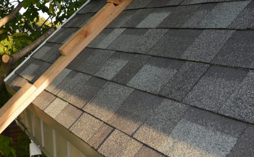 Close-up view of dark gray asphalt shingles on a residential roof with wooden ridge beam and rain gutter, showing professional roof repair and installation work