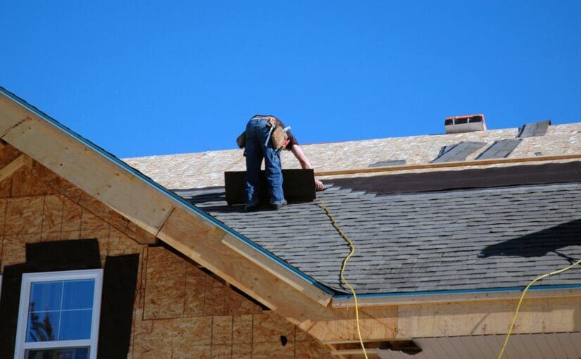 Roofer in blue shirt and harness inspecting residential roof shingles during roof assessment on partially constructed home