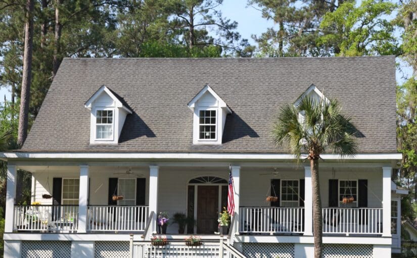 White farmhouse with gray shingle roof, two dormers, front porch with white railings, and palm trees, representing quality residential roofing and home exterior services