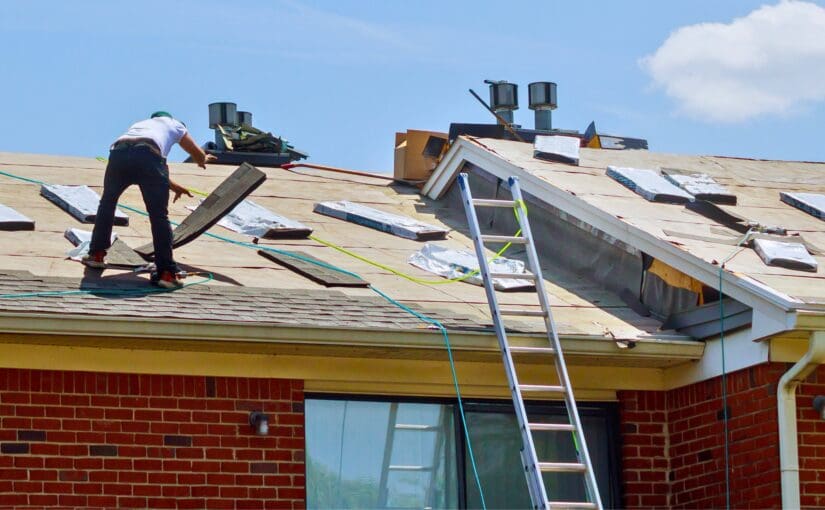 Roofer removing damaged shingles during residential roof repair on brick home with ladder and safety equipment visible