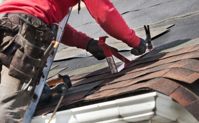 Roofer in red jacket and gloves installing asphalt shingles on residential roof with damaged brown shingles visible